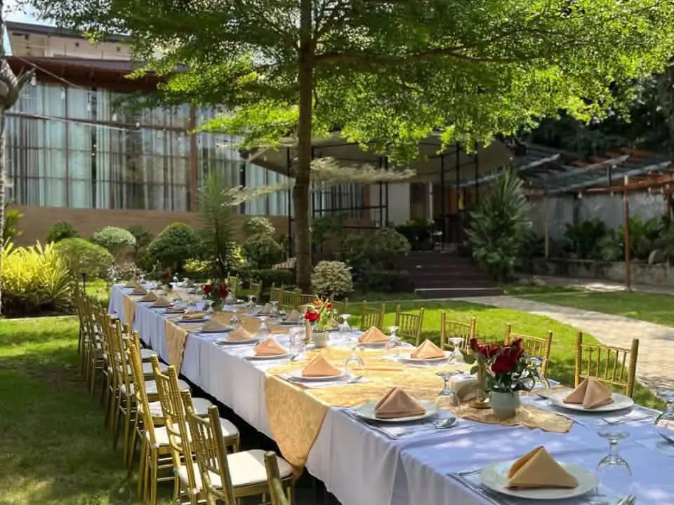 A long table laid with white cloths and gold chairs on the lawn, under the trees.