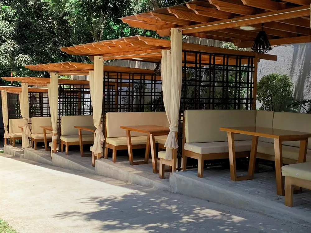 Cushioned booths and timber tables in a row under the trellis, with curtains at each post.