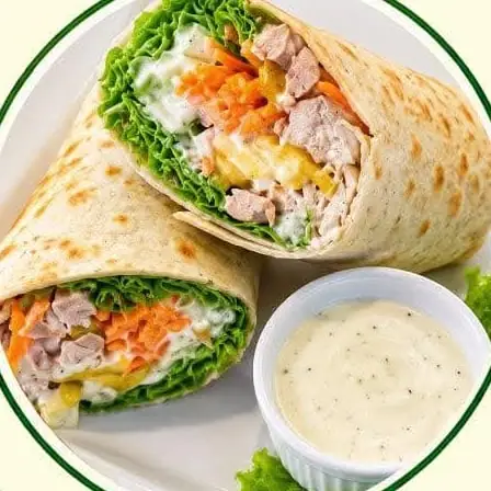 A chicken and vegetable salad wrap, cut in half, with a cup of dressing.