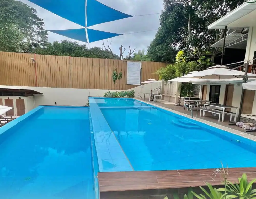 The swimming pool under blue shade sails, with umbrellas and trees around it.